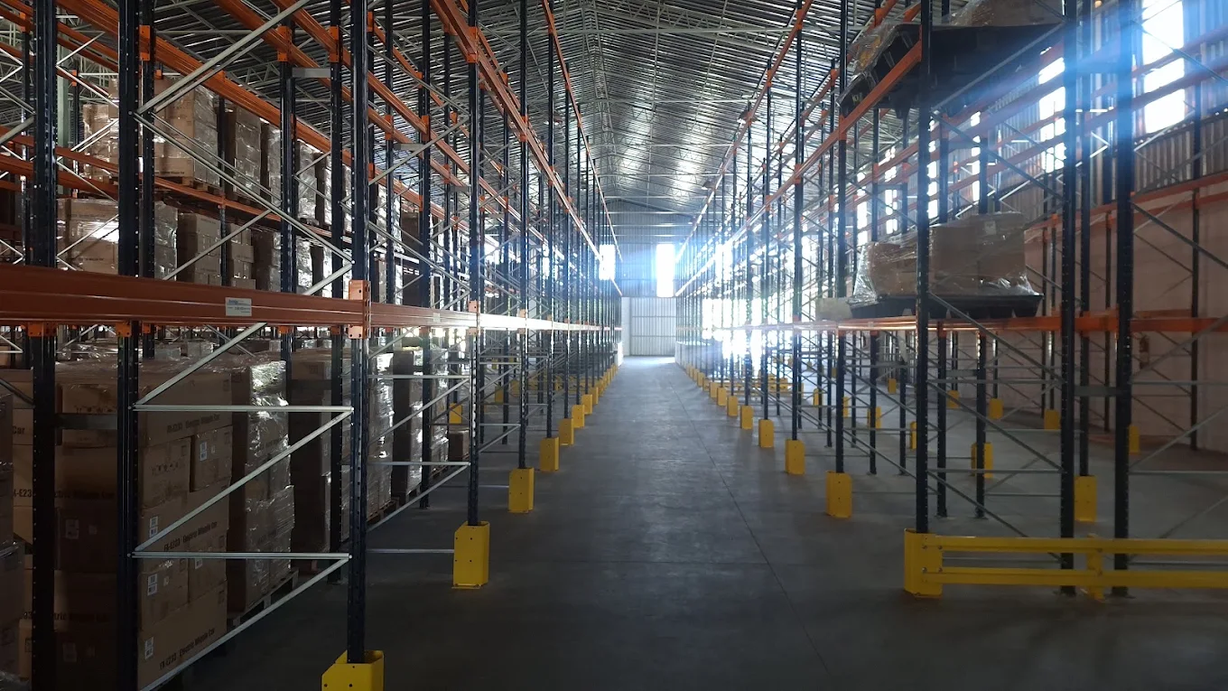 Corredor interno do armazém, com porta-paletes de vários níveis ocupados por caixas paletizadas e protetores de coluna.